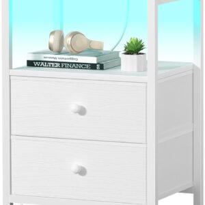 A modern white nightstand with two drawers and an open shelf illuminated by a soft blue LED light. On top of the nightstand sits a green cup and saucer beside a smartphone connected to a charging cable, highlighting the built‑in USB charging station. Inside the glowing shelf are beige headphones, two stacked books—one titled Walter Finance—and a small potted plant in a white pot. A white power cord extends from the bottom, emphasizing the nightstand’s powered design for lighting and device charging.