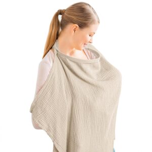 A smiling woman with blonde hair tied in a ponytail wears a light beige muslin nursing cover while breastfeeding. The soft, crinkled cotton fabric drapes over her shoulders and chest, secured around her neck with a strap that creates a small open neckline for visibility and airflow. The background is plain white, emphasizing the breathable, comfortable design of the Konssy Muslin Nursing Cover in Summer Wheat.