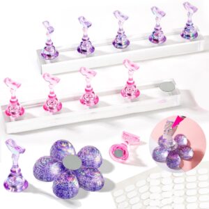 Two acrylic nail stands—one pink and one purple—each holding five magnetic holders for press‑on nails, displayed on clear rectangular bases. A glittery purple flower‑shaped base with magnets sits nearby, along with adhesive pads and a close‑up inset showing a nail being painted on the magnetic holder.