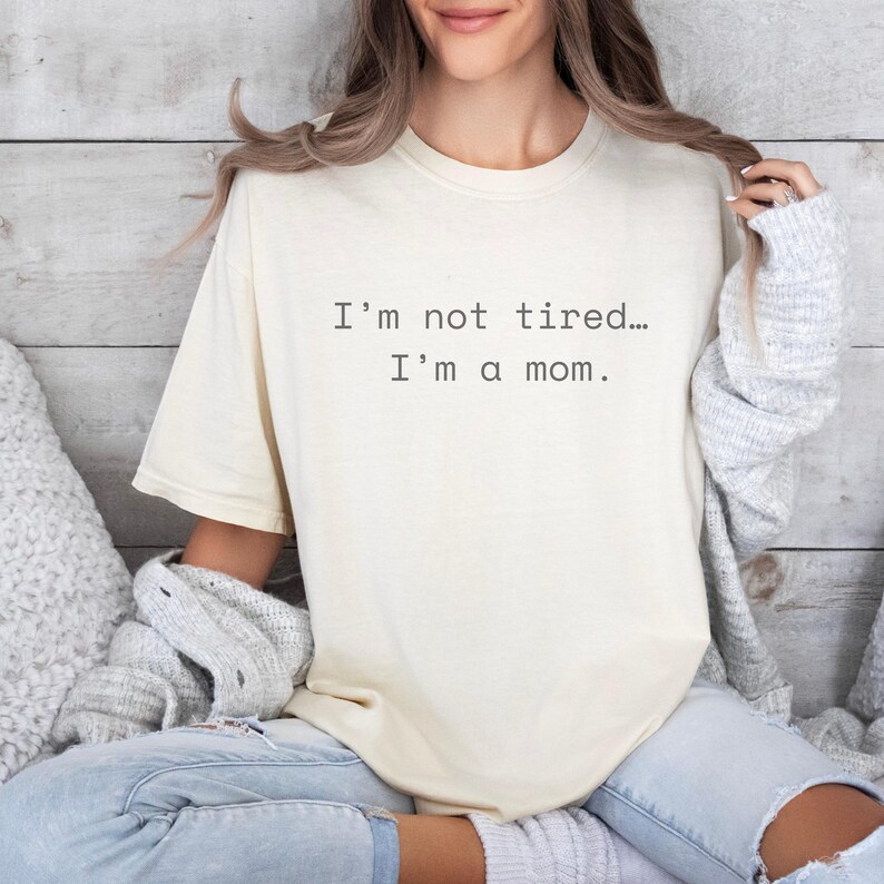 A cream not tired mom shirt featuring minimalist black text that reads “I’m not tired… I’m a mom,” styled with a cozy gray cardigan and light ripped jeans in a relaxed home setting.