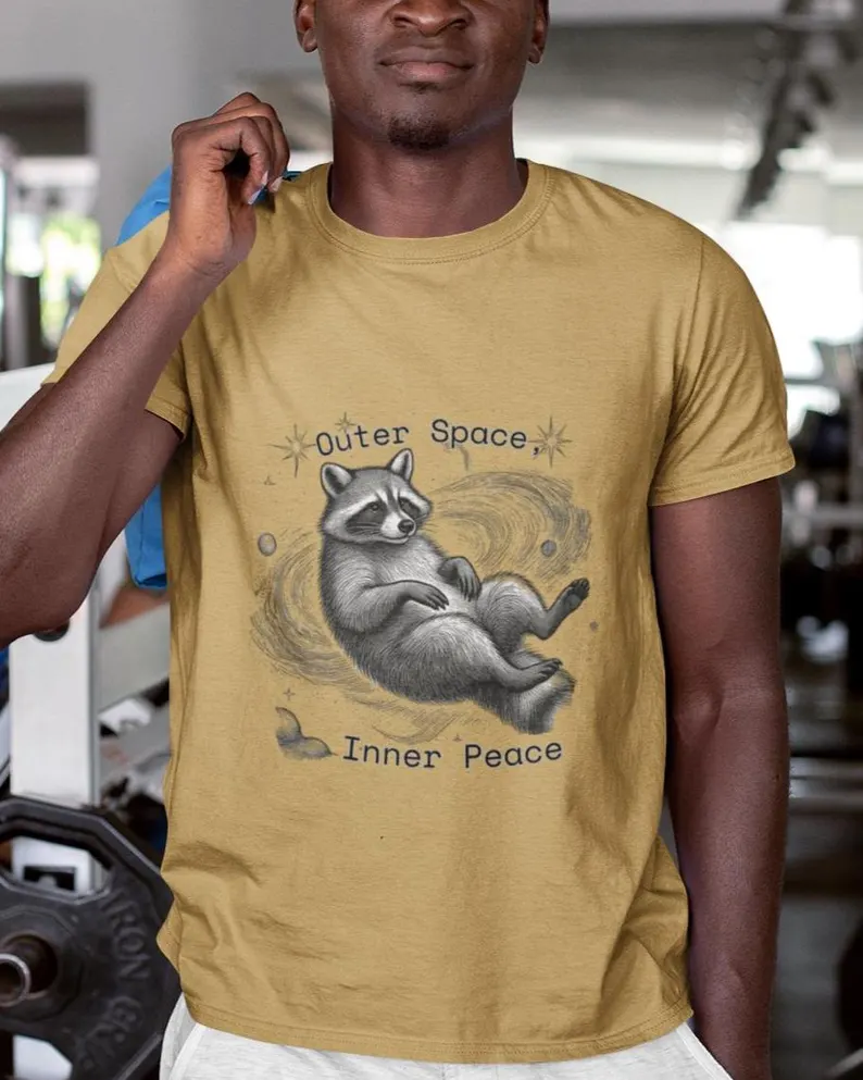A tan outer space inner peace shirt featuring a raccoon floating peacefully in a galaxy with stars and planets, and the text “Outer Space, Inner Peace,” styled in a gym setting.