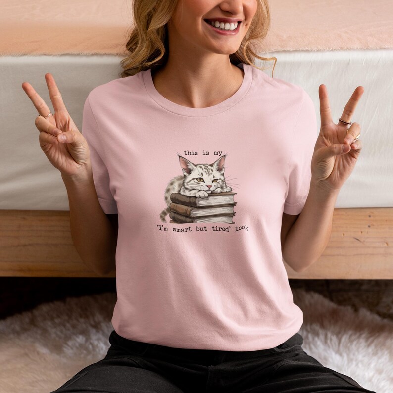 A light pink smart but tired shirt featuring an illustration of a sleepy cat resting on books with the text “This is my ‘I’m smart but tired’ look,” styled casually indoors.