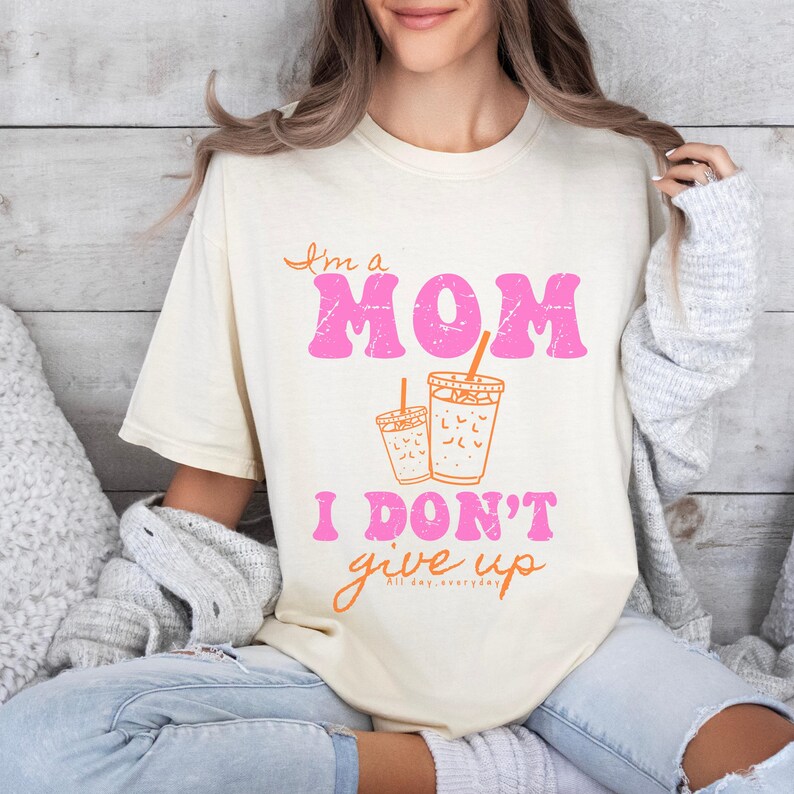 A cream mom don’t give up shirt featuring pink and orange text with two illustrated coffee cups, styled with cozy gray cardigan and jeans in a relaxed home setting.