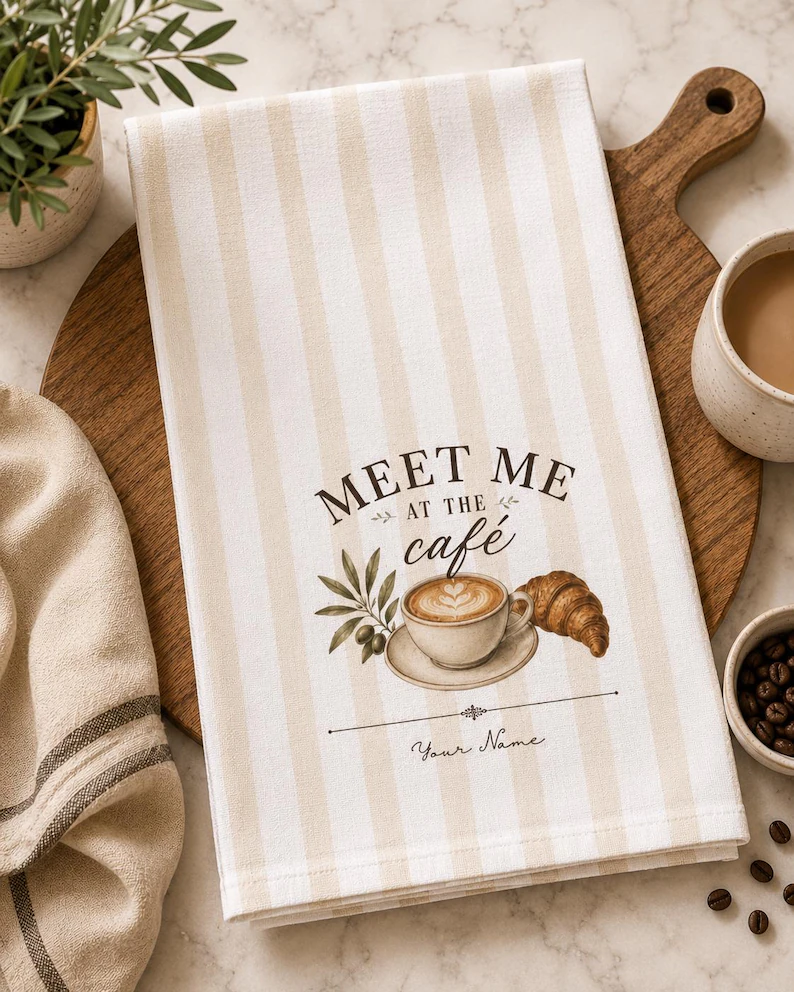 A beige and white striped meet me cafe towel featuring a latte, croissant, and olive branch design with personalized name text, styled on a wooden cutting board beside coffee and kitchen accessories.