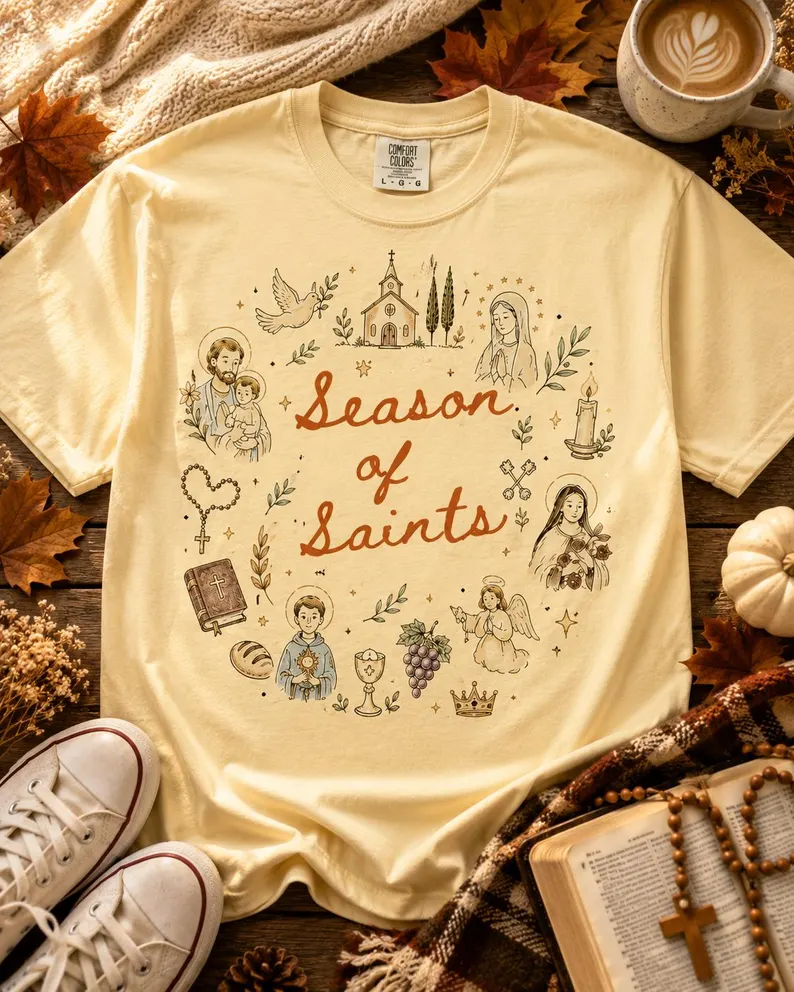 A cream season of saints shirt featuring vintage Catholic illustrations of saints, a church, and faith symbols with the text “Season of Saints,” styled with fall leaves, coffee, and a rosary.