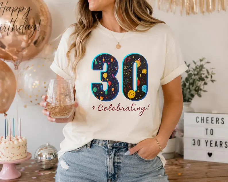 A cream celebrating 30 shirt featuring colorful party illustrations inside the number “30” with fireworks, balloons, and confetti, styled with jeans and birthday decorations.