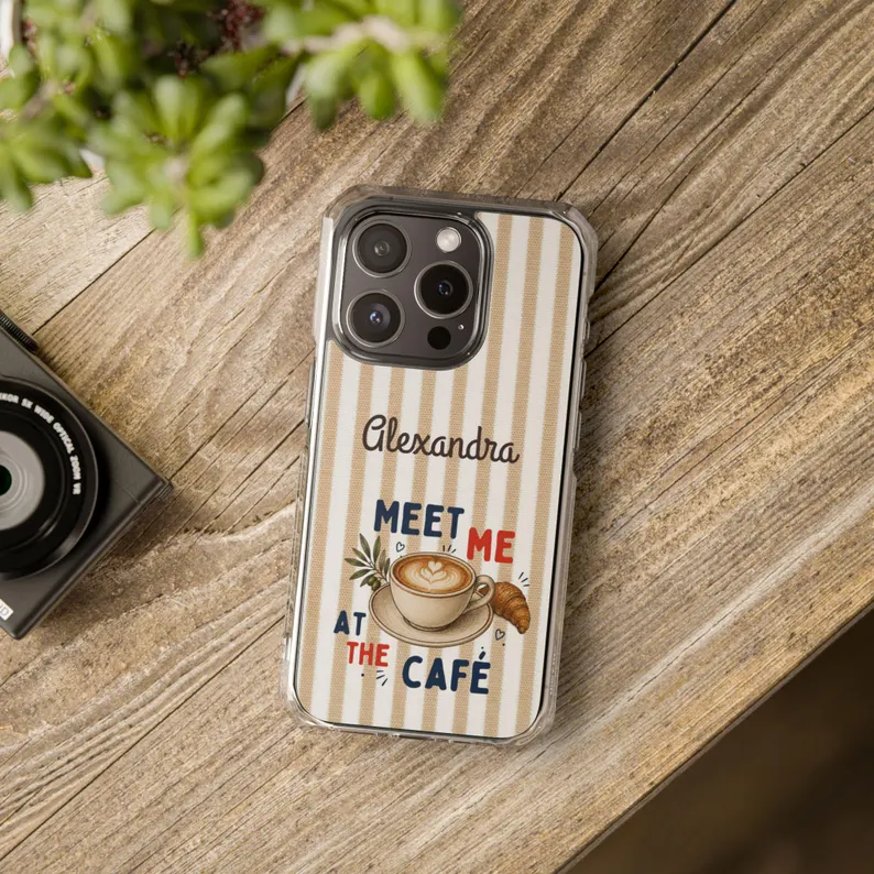A beige and white striped meet me cafe case featuring a latte, croissant, and olive branch design with personalized name text, styled on a wooden table beside a camera and plant.