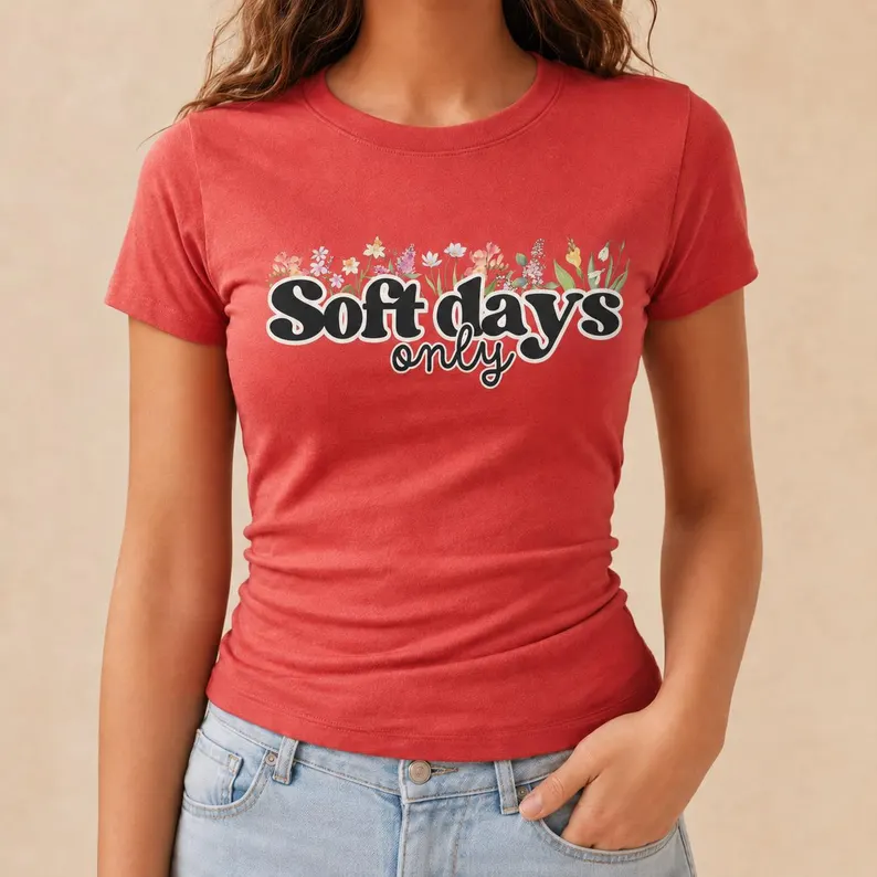 A woman wearing a red soft days shirt with floral illustrations and bold text “Soft Days Only,” styled casually with light denim jeans in a neutral setting.