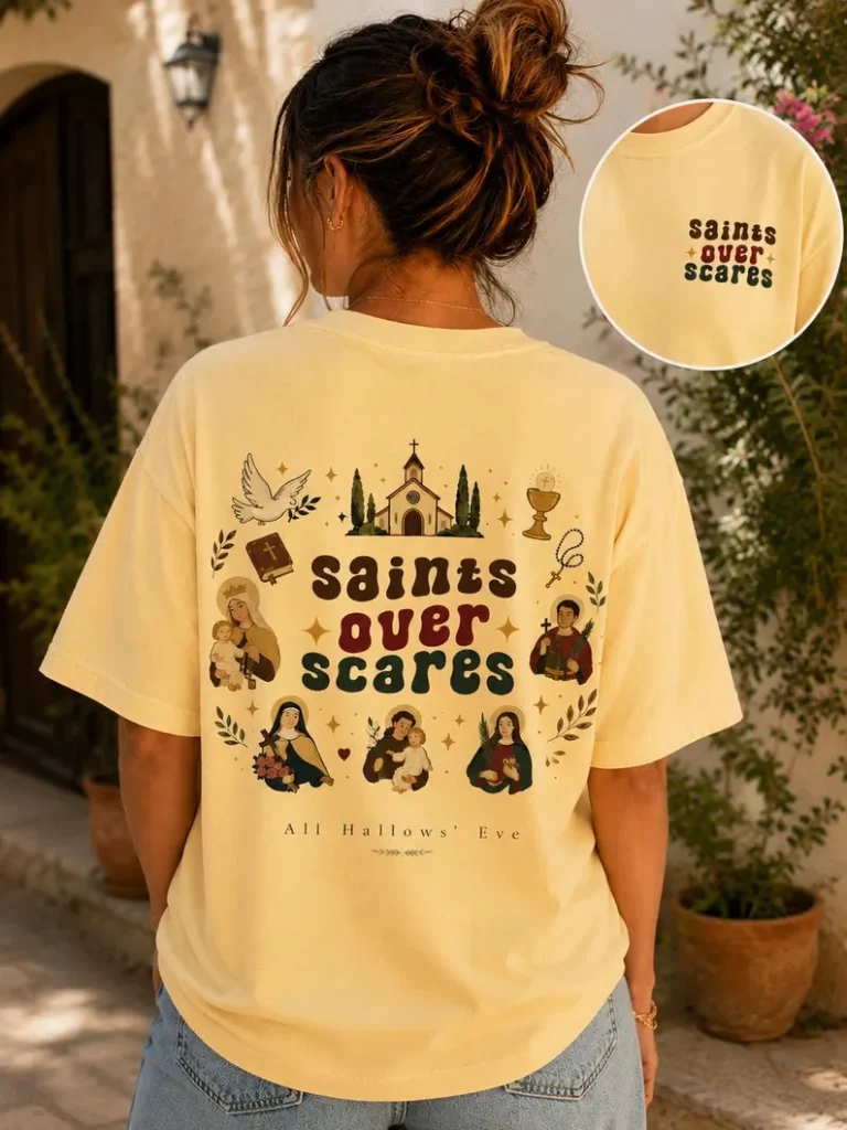 A yellow saints over scares shirt featuring vintage Catholic illustrations of saints, a church, and faith symbols with the text “Saints Over Scares – All Hallows’ Eve,” styled outdoors with fall décor.