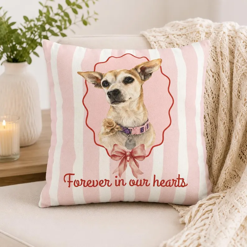 A pink and white striped pet memorial pillow featuring a portrait of a small dog with a red bow and the text “Forever in our hearts,” styled on a beige sofa with a candle and cozy blanket.