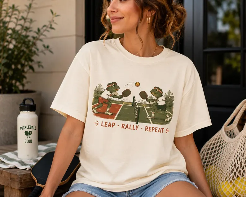 A cream pickleball frog shirt featuring two cartoon frogs playing pickleball on a court with paddles and a ball, and the text “Leap • Rally • Repeat,” styled with shorts and a water bottle.